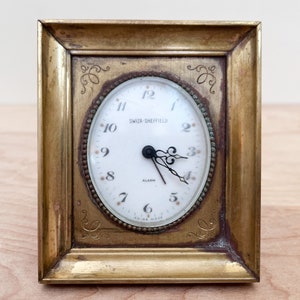 May include: A gold-toned brass framed alarm clock with a white face and black hands. The clock face reads "SWIZA-SHEFFIELD" and "ALARM".