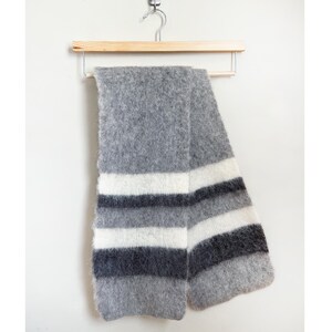 May include: A gray scarf with horizontal stripes of white, dark gray, and medium gray. The scarf is made of a soft, textured material and is hanging on a wooden hanger against a white background. The scarf is a winter accessory.