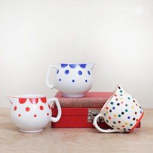 May include: Three ceramic pitchers with white backgrounds. One pitcher has red polka dots, another has blue polka dots, and the third has a pattern of colorful squares. The pitchers are on a wooden surface.