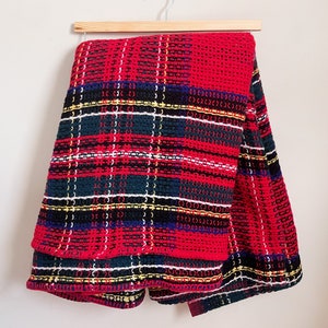 May include: Red, green, black, and white plaid throw blanket with a fringed edge. The blanket is folded and hanging on a wooden hanger.