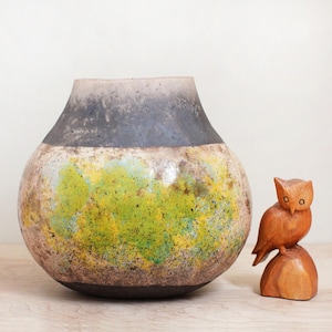 May include: A large, round, ceramic vase with a black, white, and green glaze. The vase has a wide mouth and a rounded bottom. It is sitting on a wooden surface next to a small, wooden owl figurine.