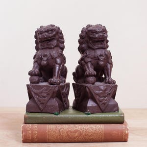 May include: Two dark brown Foo Dog statues, each perched on a geometric base, sit atop a stack of vintage books. The statues have detailed manes and are holding a ball under their paws. The books have green and brown covers.
