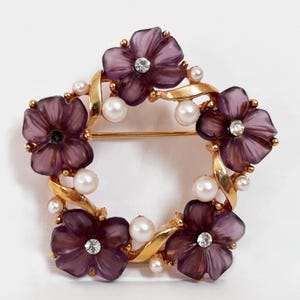 May include: A gold tone brooch with a circular design featuring four purple flowers with rhinestones and white pearl accents.
