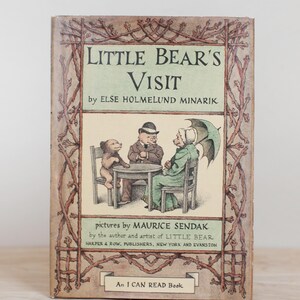 May include: Vintage children's book titled "Little Bear's Visit" by Else Holmelund Minarik, with illustrations by Maurice Sendak. The cover features a charming illustration of three bears seated around a table. The book is framed by a decorative border of brown branches.