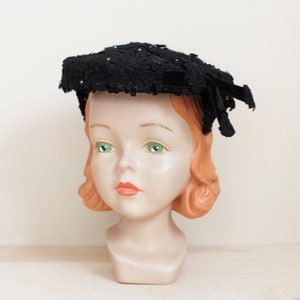 May include: A black lace hat with a velvet band and small pink beads. The hat is on a mannequin head with red hair.