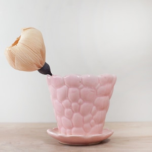 May include: A light pink ceramic flower pot with a textured, bubble-like pattern. The pot has a matching saucer and holds a dried, beige flower with a dark stem. The pot is on a wooden surface.
