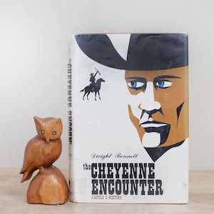 May include: A vintage book titled "The Cheyenne Encounter" by Dwight Bennett, featuring a cowboy illustration. Beside the book is a carved wooden owl figurine.