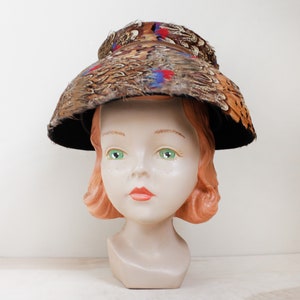 May include: A vintage hat made of brown and black feathers with red and blue accents. The hat is perched on a mannequin head.