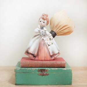 May include: A porcelain figurine of a girl in a white and pink dress holding a dried corn husk. The figurine is sitting on top of two vintage books and a green wooden box.