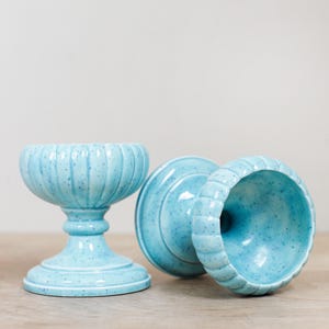 May include: Three light blue ceramic pedestal bowls with a speckled finish. One bowl is upright, while the other two are on their sides. The bowls have a ribbed design around the rim and a pedestal base.
