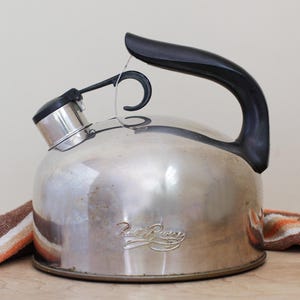 May include: A stainless steel tea kettle with a black handle and spout. The kettle has a rounded body and a black lid. The brand name "Paul Revere" is etched on the side. A striped towel is partially visible.