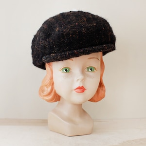 May include: A black tweed newsboy cap with a brown fleck pattern. The cap is on a mannequin head with red hair.