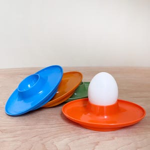 May include: A white egg sits in an orange egg cup, with three other colorful egg cups in blue, brown, and green. The egg cups are made of plastic and have a saucer-like base.