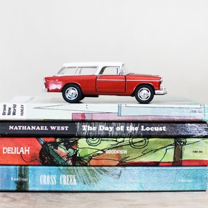 May include: A stack of four paperback books with a red toy station wagon on top. The books are titled "Brave New World", "The Day of the Locust", "Delilah", and "Cross Creek".