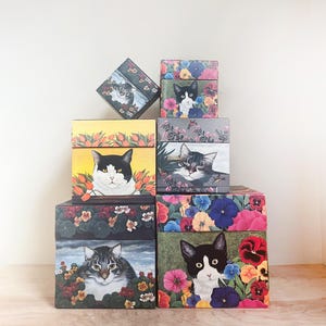 May include: A collection of decorative boxes featuring cat portraits surrounded by colorful flowers. The boxes showcase various cat breeds with different floral arrangements, including tulips, pansies, and other blooms. The boxes are stacked in a pyramid shape.