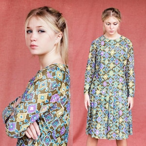 May include: A vintage long-sleeve dress with a geometric pattern in olive green, purple, blue, and yellow. The dress has a round neckline and a knee-length hemline. The model is wearing the dress in a studio setting.