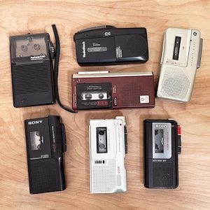 Puede incluir: Una colección de grabadoras de cassette vintage en varios colores, incluyendo negro, blanco y burdeos. Las grabadoras presentan marcas como Sony, RadioShack y GE. Algunas tienen cintas de cassette y botones de control visibles. Las grabadoras están dispuestas sobre una superficie de madera clara.
