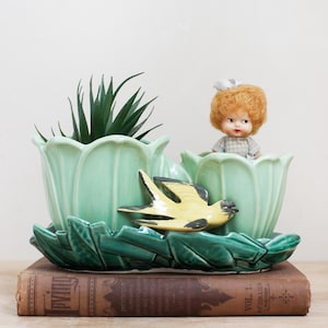 May include: A vintage ceramic planter in the shape of a tulip, light green in color, with a dark green leaf-shaped base. A yellow and black bird figurine rests on the planter. A small doll and a faux plant are inside.