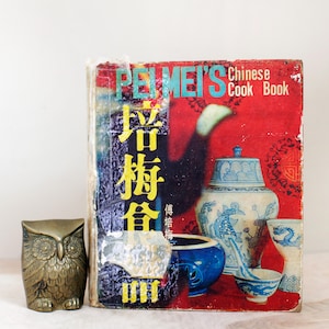 May include: A vintage cookbook titled "Pei Mei's Chinese Cook Book" with a red cover and a colorful illustration of Chinese porcelain. The title is in blue and yellow lettering. The book is next to a brass owl figurine.