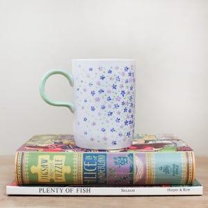 May include: A white ceramic mug with a light green handle, decorated with small blue and purple floral patterns. The mug rests on a stack of books, including "Alice in Wonderland" and "Plenty of Fish".