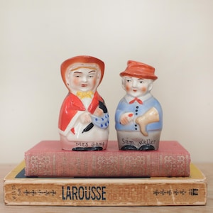 May include: A pair of vintage ceramic salt and pepper shakers depicting Mrs. Gamp and Sam Weller. The shakers are painted in red, white, blue, and yellow. The shakers are sitting on top of a red book with the word "LAROUSSE" printed on the cover.