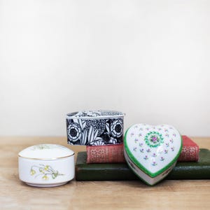 May include: A collection of decorative ceramic boxes in various shapes and designs. Includes a heart-shaped box with floral accents, a rectangular box with a black and white floral pattern, and a round box with a floral design.
