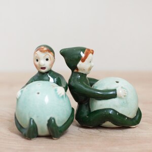 May include: A pair of vintage ceramic salt and pepper shakers shaped like elves holding green balls. The elves are wearing green hats and have brown hair.