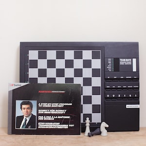 May include: A black Saitek Kasparov Team-Mate electronic chess computer with a gray and black checkered board. The device features a control panel with buttons and a display. A matching instructional booklet and two chess pieces are included.