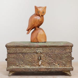 May include: A wooden owl figurine perched atop a carved wooden base, placed on a decorative metal box. The box has an embossed floral design and a latch closure. The owl is light brown, and the box has a bronze finish.