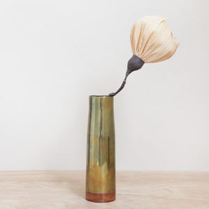 May include: A tall, brass-colored vase with a copper base holds a unique, handmade flower. The flower has a dark stem and a large, light-colored, textured bloom. The vase is cylindrical and sits on a light-colored wooden surface.