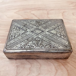 May include: A rectangular silver box with intricate engraved floral and geometric patterns on the lid. The box has a hinged design and a slightly tarnished appearance, suggesting age. The detailed carvings create a textured surface.