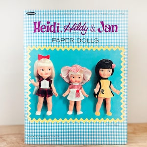 May include: A vintage paper doll book titled "Heidi, Hildy & Jan" with three dolls on the cover. The cover has a blue and white checkered pattern. The dolls are wearing different outfits and have different hairstyles. The text "No Scissors Necessary Clothes Just Press Out" is at the top.