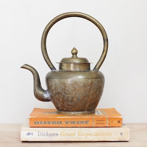 May include: Antique brass teapot with a curved handle and floral engravings. The teapot sits atop two vintage books, "Oliver Twist" and "Great Expectations". The books are orange and cream colored.