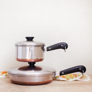 May include: A stainless steel saucepan with a copper base and a black handle sits stacked atop a stainless steel skillet with a copper base and a black handle. Both pieces feature a matching black lid knob and a metal hanging loop on the handle.