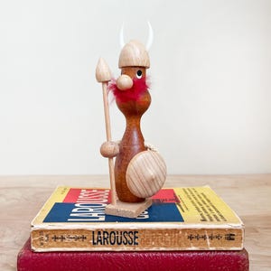 May include: A wooden Viking figurine with a spear and shield stands atop two vintage books. The figurine has a red beard, a wooden helmet with white horns, and a round wooden shield. The books are red and yellow.