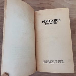 Op de afbeelding: Een open boek met verouderde, cr&egrave;mekleurige pagina's. De titel "PERSUASION" van Jane Austen is gedrukt in zwarte, vette letters. Het boek is uitgegeven door Magnum Easy Eye Books, Lancer Books, New York.