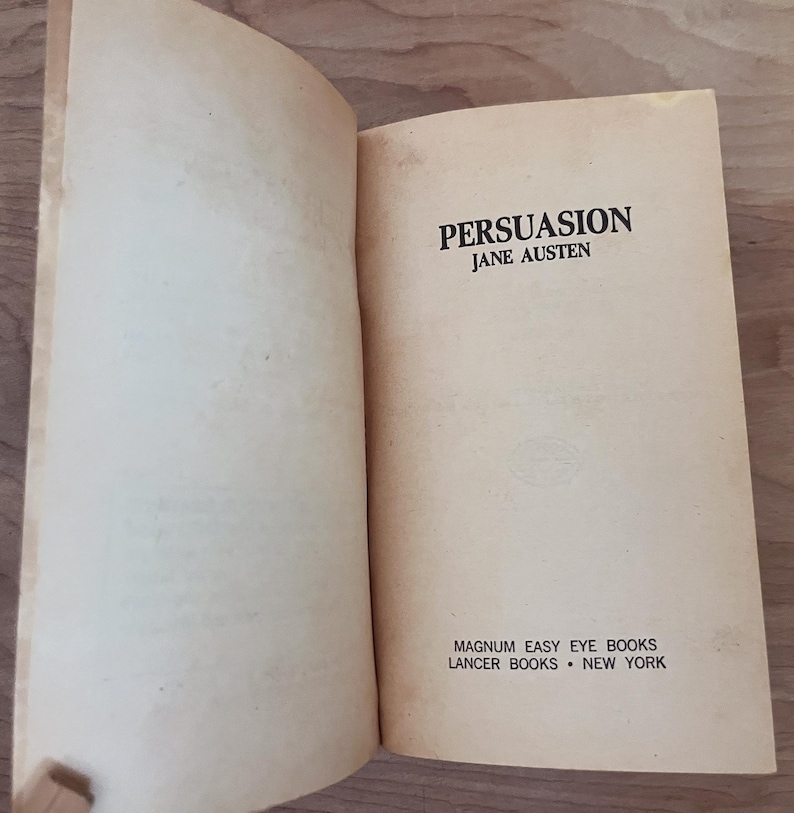 Op de afbeelding: Een open boek met verouderde, cr&egrave;mekleurige pagina's. De titel "PERSUASION" van Jane Austen is gedrukt in zwarte, vette letters. Het boek is uitgegeven door Magnum Easy Eye Books, Lancer Books, New York.