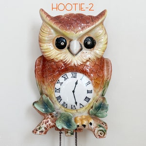 May include: A brown and yellow ceramic owl clock with black eyes and a white clock face with black Roman numerals. The owl is perched on a branch with green leaves.