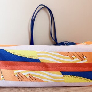 May include: A large orange tote bag with a blue and white striped pattern and a yellow rope design. The bag has two blue handles.