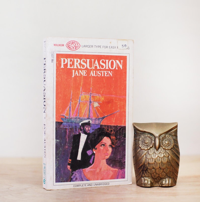 Op de afbeelding: Een vintage paperback boek getiteld "Persuasion" van Jane Austen, met een omslagillustratie van een schip en figuren. Daarnaast een messing uil beeldje, een decoratief item.
