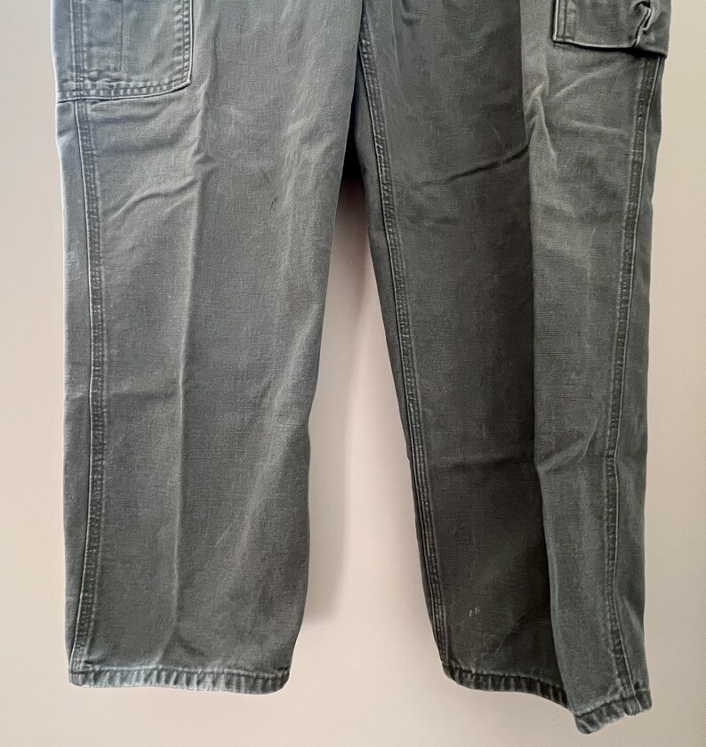 Może przedstawiać: Spodnie robocze w ciemnozielonym kolorze z dwoma dużymi kieszeniami z przodu i dwoma dużymi kieszeniami z tyłu. Spodnie mają prosty kr&oacute;j i luźny fason.