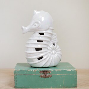 May include: White ceramic seahorse figurine with a pearlescent finish. The seahorse is standing on a teal green wooden box.