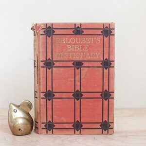 May include: A vintage red book with a black and white geometric pattern on the cover. The book is titled "Peloubet's Bible Dictionary". A small gold metal mouse figurine is in front of the book.