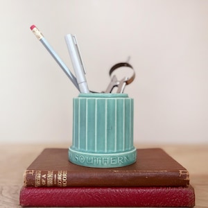 May include: A teal ceramic desk organizer with vertical grooves, holding a pencil, pen, and scissors. The organizer sits atop two stacked books with brown and red covers. The text "SOUTHERN" is visible on the organizer.