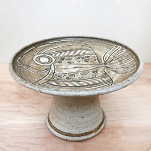 May include: A white ceramic cake stand with a brown stylized fish design. The stand has a round top and a pedestal base.