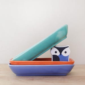 May include: A stack of three rectangular ceramic dishes in teal, orange, and blue. A small, decorative owl figurine with blue and black details sits between the orange and teal dishes. The items are arranged on a wooden surface.
