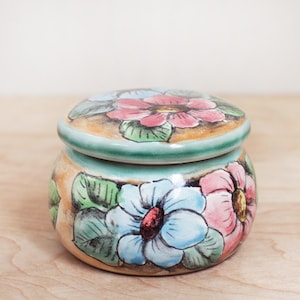 May include: A small, round ceramic jar with a lid, adorned with hand-painted floral designs. The jar features a turquoise rim and lid, with flowers in shades of pink, blue, and yellow. The background is a light beige color.