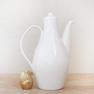 May include: A tall, white ceramic pitcher with a curved handle and spout, next to a small, gold-colored mouse figurine. The pitcher has a smooth, glossy finish. The mouse figurine is detailed with small ears and a rounded body.