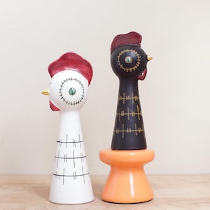 May include: Two ceramic rooster figurines. One is white with a red comb, green eye, and black cross-stitch details. The other is black with a red comb, green eye, and gold cross-stitch details, set on an orange base. Both have gold beaks.