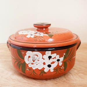 May include: A round, orange ceramic casserole dish with a lid. The dish is decorated with white floral designs and green leaves. It has a dark brown rim and handles. The lid has a small, round handle.